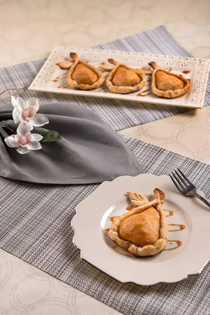A dessert plate with a beautiful Carmel Pear Pie. A folded napkin and a tray of 3 Carmel Pear Pies. All have been treated to limit browning of the fruit.