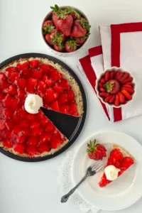 An overhead view of a table with a round strawberry pizza on a pizza pan, slice of pie, and sliced strawberries