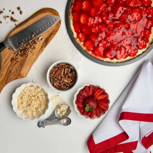 Strawberry Pizza dessert, sitting on a white table, with these ingredients in small bowls crumble crust, sliced strawberries, nuts and a small organic chopping block with a knife and chopped nuts.