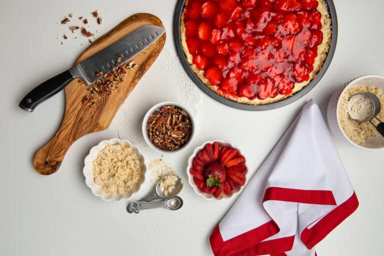 Strawberry Pizza dessert, sitting on a white table, with these ingredients in small bowls crumble crust, sliced strawberries, nuts and a small organic chopping block with a knife and chopped nuts.