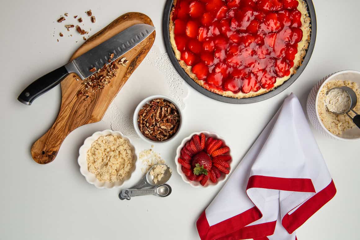 Strawberry Pizza dessert, sitting on a white table, with these ingredients in small bowls crumble crust, sliced strawberries, nuts and a small organic chopping block with a knife and chopped nuts.