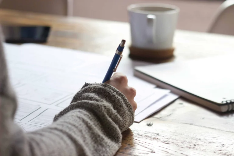 Journaling Techniques for a better you a woman sitting at a table writing in a journal. also on the table are notebooks, coffee cup and papers