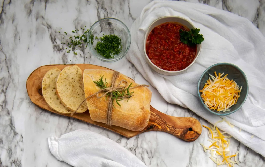 A white marble table top sitting on top is a white kitchen towel, small bowl of cheddar cheese, small bowl of chopped parsley, small wooden cutting board with sliced French bread and a bowl of red Spaghetti Sauce.