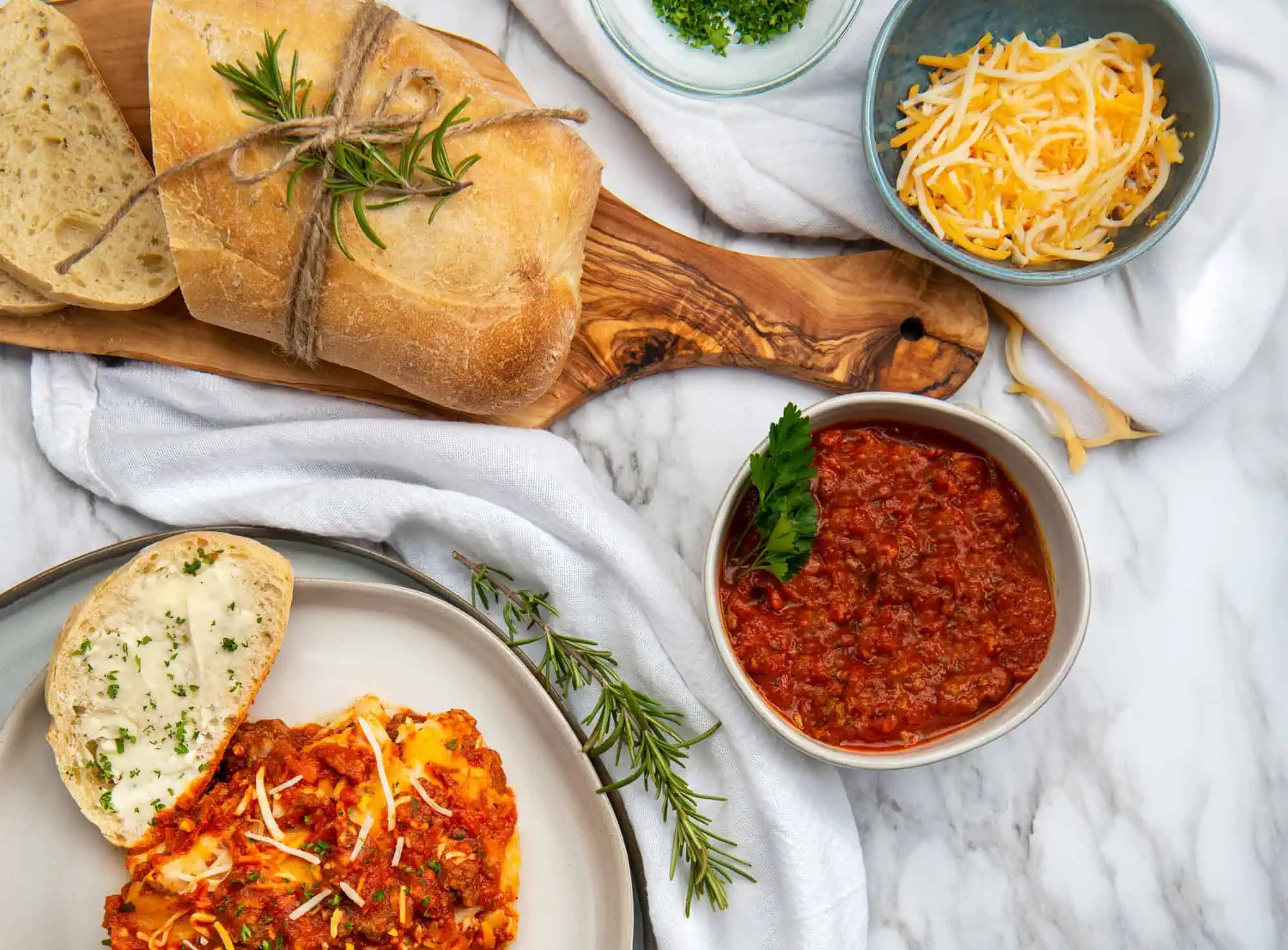Semi-Homemade Spaghetti Sauce. A white marble table top sitting on top is a white kitchen towel, small bowl of cheddar cheese, small bowl of chopped parsley, small wooden cutting board with sliced French bread and a bowl of red Spaghetti Sauce. Also, a plate with lasagna and French bread.
