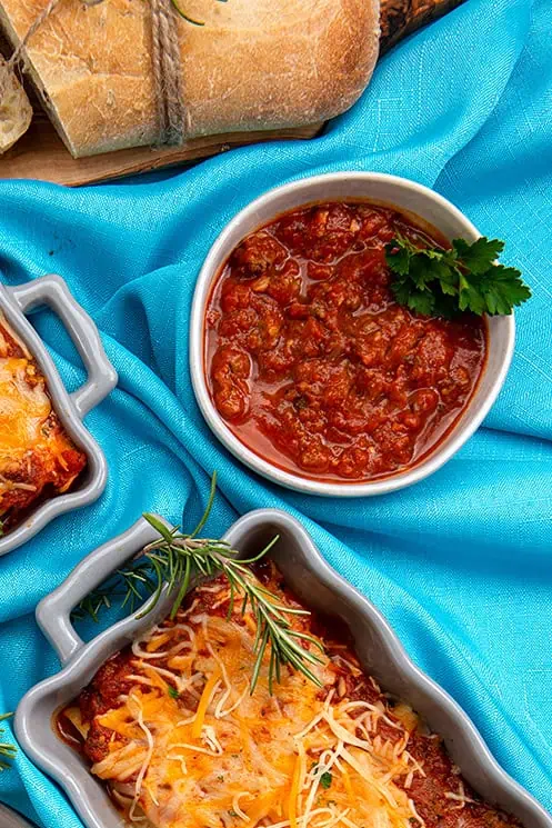 Table setting with a turquoise table cloth, setting on the table is a bowl of spaghetti sauce and a serving of lasagna.