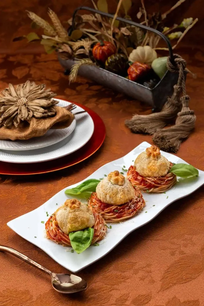 Delectable & Delicious Meatball Wellington An up close and side view of a platter of spaghetti topped with meatball wellington. In the background and slightly out-of-focus dinner plate and centerpiece.