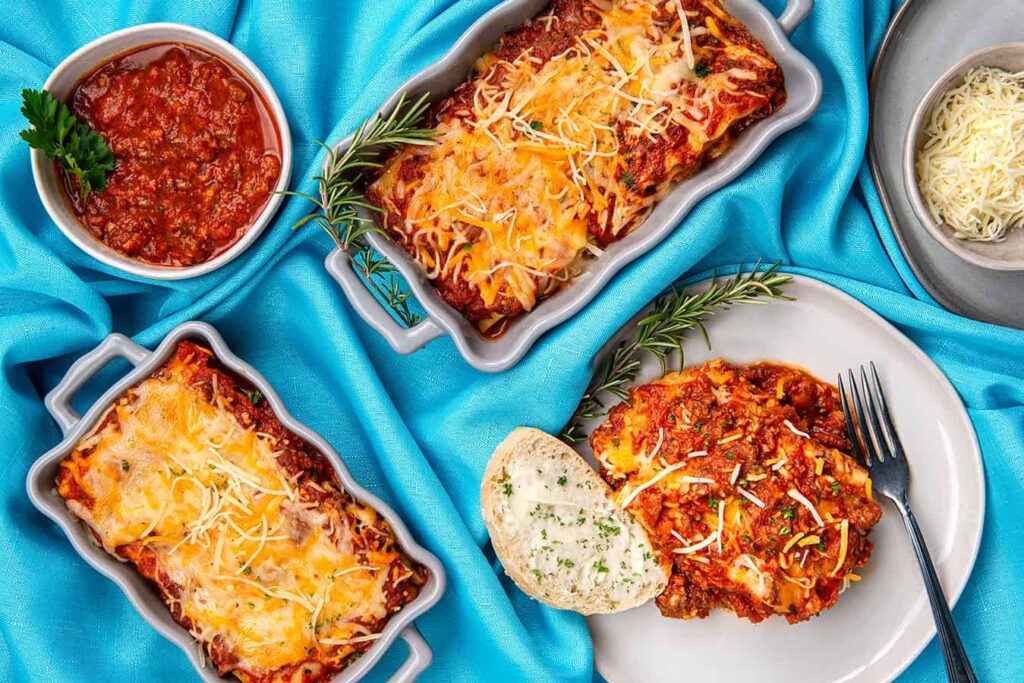 Irresistible Layers of Lasagna You’ll Love to Make and Share. A close up overhead shot of a slice of lasagna sitting in a dinner plate with French bread. Two different sized trays of cooked lasagna, small bowls of Semi Home-made Spaghetti Sauce and grated cheese.