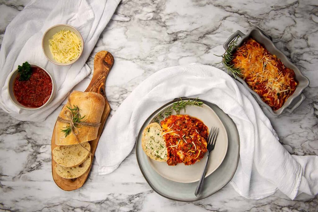 Irresistible Layers of Lasagna You’ll Love to Make and Share. A close up overhead shot of a slice of lasagna sitting in a dinner plate with French bread. A loaf of French Bread, tray of lasagna, small bowls of grated cheese, and a bowl of Semi Home-made Spaghetti Sauce.