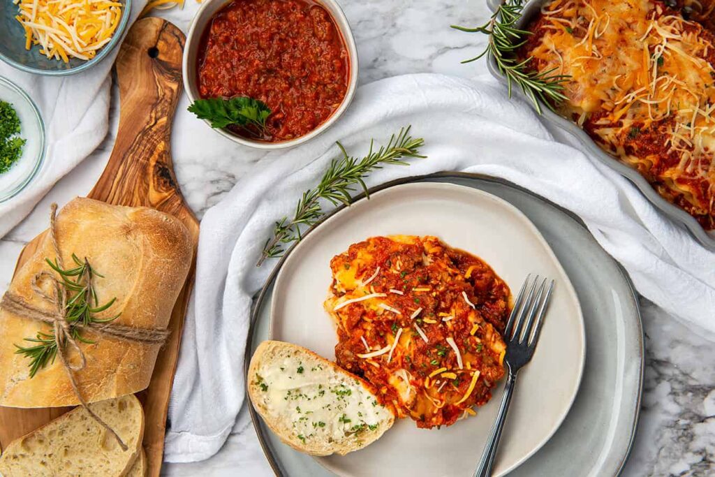 Irresistible Layers of Lasagna You’ll Love to Make and Share. A close up overhead photo of a slice of lasagna sitting in a dinner plate with French bread. Also in the photo and a bowl of Semi Home-made Spaghetti Sauce, tray of lasagna, grated cheese and chopped parsley.