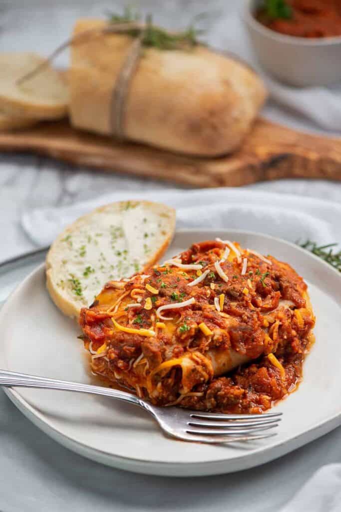 Irresistible Layers of Lasagna You’ll Love to Make and Share. A close up photo of a slice of lasagna sitting in a dinner plate with French bread. Out of focus in the background is a loaf of French Bread and a bowl of Semi Home-made Spaghetti Sauce.