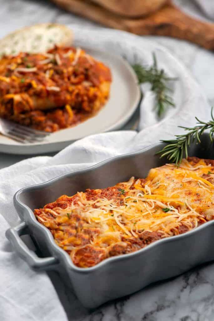 Irresistible Layers of Lasagna You’ll Love to Make and Share. A close up photo of a baking dish of lasagna sitting in a dinner plate with French bread. Out of focus in the background is a loaf of French Bread and plate of lasagna.
