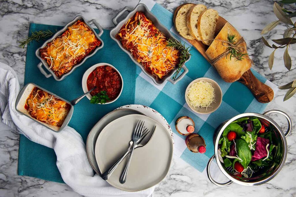 Irresistible Layers of Lasagna You’ll Love to Make and Share. A close up overhead shot of a slice of lasagna sitting in a dinner plate with French bread. Three different sized trays of cooked lasagna, small bowls of Semi Home-made Spaghetti Sauce, grated cheese, salt and pepper shakers, colander of lettuce and a partially sliced loaf of French bread.