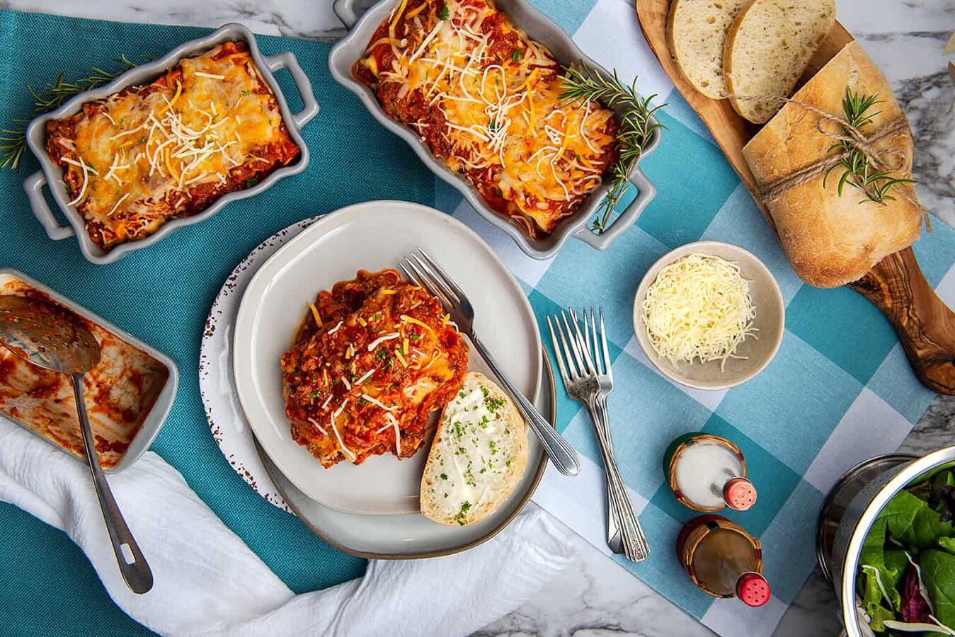 Irresistible Layers of Lasagna You’ll Love to Make and Share. A close up overhead shot of a slice of lasagna sitting in a dinner plate with French bread. Two different sized trays of cooked lasagna, small bowls of Semi Home-made Spaghetti Sauce, grated cheese and a partially sliced loaf of French bread.