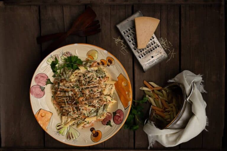 Creamy Chicken Pasta A Dreamy, Comforting Delight blog post. In this image there is a large bowl filled with pasta and chicken, nearby is a cheese grater with a large triangle of Parmesan cheese. In the right hand corner is a colander of cooked pasta