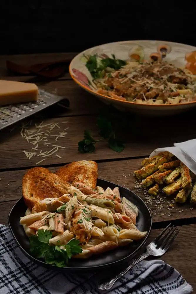 Creamy Chicken Pasta A Dreamy, Comforting Delight blog post. In this image there is a large bowl filled with pasta and chicken, nearby is a cheese grater with a large triangle of Parmesan cheese. In the right hand corner is a colander of cooked pasta