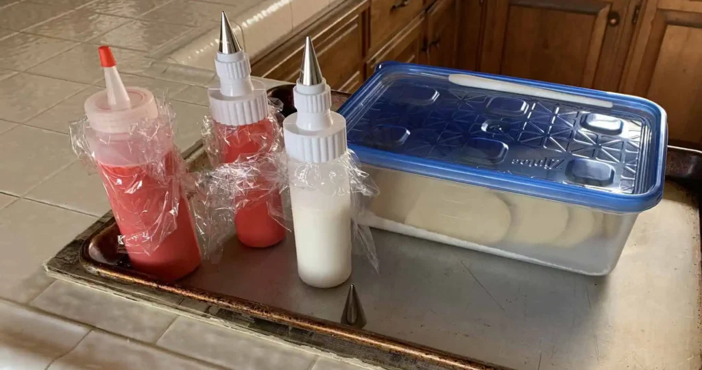 IMG_2464 Cookie decorating setup: two bottles of red icing and one bottle of white icing, all covered with plastic wrap and fitted with piping tips, sit on a baking sheet next to a container of undecorated cookies with a blue lid.