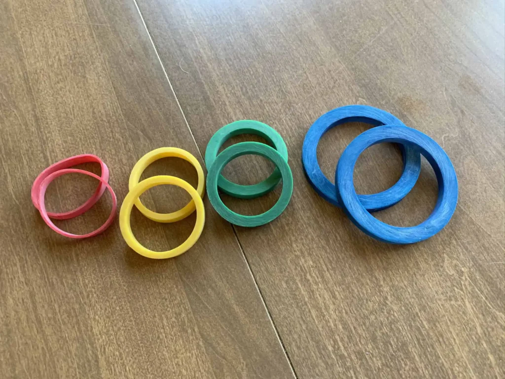 IMG_2555 Four pairs of rolling pin spacer rings in different colors and sizes, arranged on a wooden surface. The colors, from left to right, are red, yellow, green, and blue, each representing a different thickness for rolling out dough evenly.