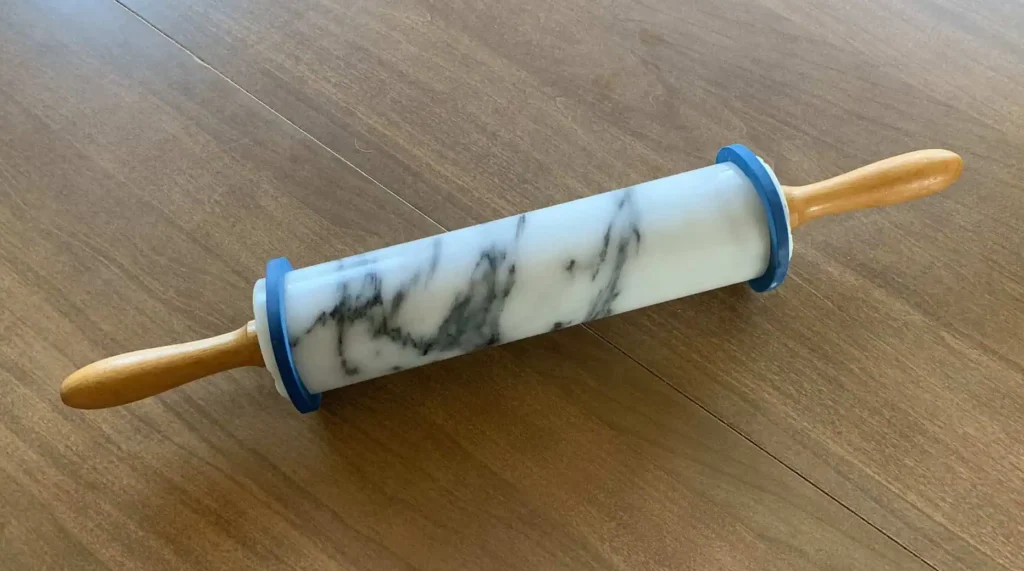 IMG_2556 A marble rolling pin with wooden handles and blue spacer rings attached to each end, resting on a wooden surface. The spacer rings help ensure an even dough thickness while rolling.