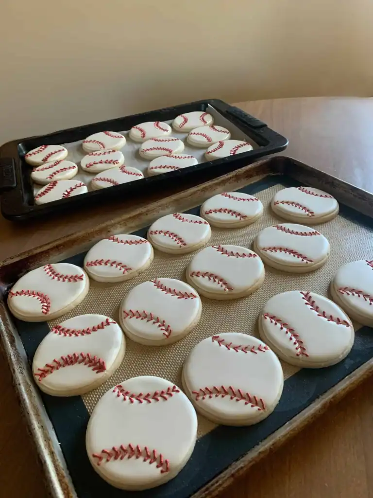 Two trays of decorated baseball sugar cookies with red royal icing stitching on a wood table