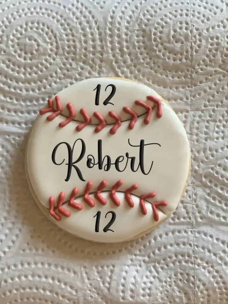 Round sugar cookie decorated like a baseball with red stitching, the name Robert, and the number 12