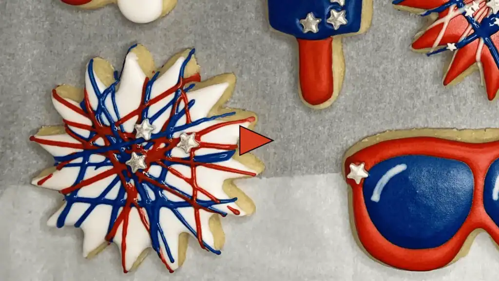 A decorated firework sugar cookie with a white royal icing base, drizzled with red and blue icing and topped with silver star sprinkles. Video play icon overlayed as a placeholder image.
