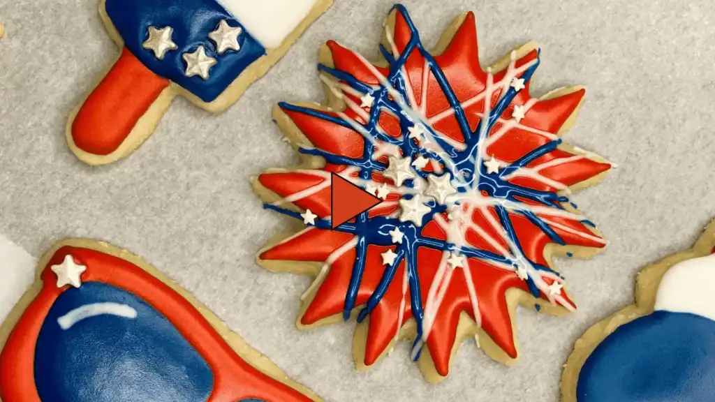 A decorated firework sugar cookie with a red royal icing base, drizzled with blue and white icing and topped with silver and white star sprinkles. Video play icon overlayed as a placeholder image.