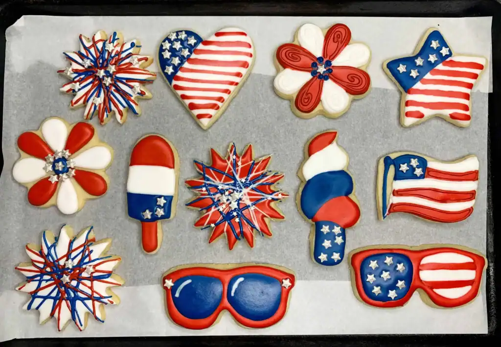 A full tray of 4th of July sugar cookies decorated with royal icing in red, white, and blue. Designs include three firework cookies, two flower cookies, a heart flag, a waving flag, a popsicle, a frosty cone, a star flag, and two pairs of patriotic sunglasses.