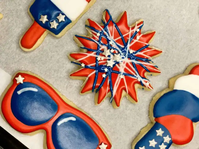 A close-up of a red firework sugar cookie decorated with blue and white icing drizzles and silver stars, surrounded by partial views of a frosty cone, red-white-blue sunglasses, and a patriotic popsicle cookie.