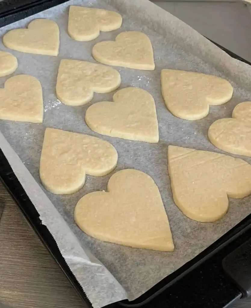 Freshly baked heart-shaped sugar cookies cooling on a parchment-lined baking sheet, resting on a stovetop. The cookies are lightly golden and ready for decorating.