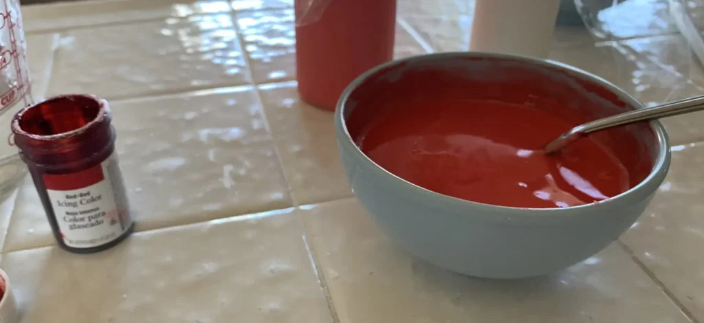 A small bowl filled with vibrant red royal icing sits on a tiled counter with a spoon, next to a container of "Red-Red" icing gel color and a measuring cup. The setup shows the process of coloring royal icing red for cookie decorating.