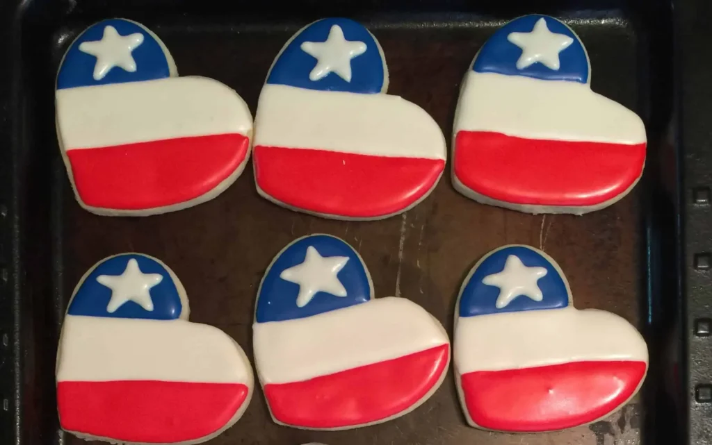 Heart-shaped sugar cookies decorated with red, white, and blue icing in American flag-inspired designs. Some feature a single white star, others have red and white stripes with clusters of small hearts or dots in the blue sections, all arranged on a baking tray.