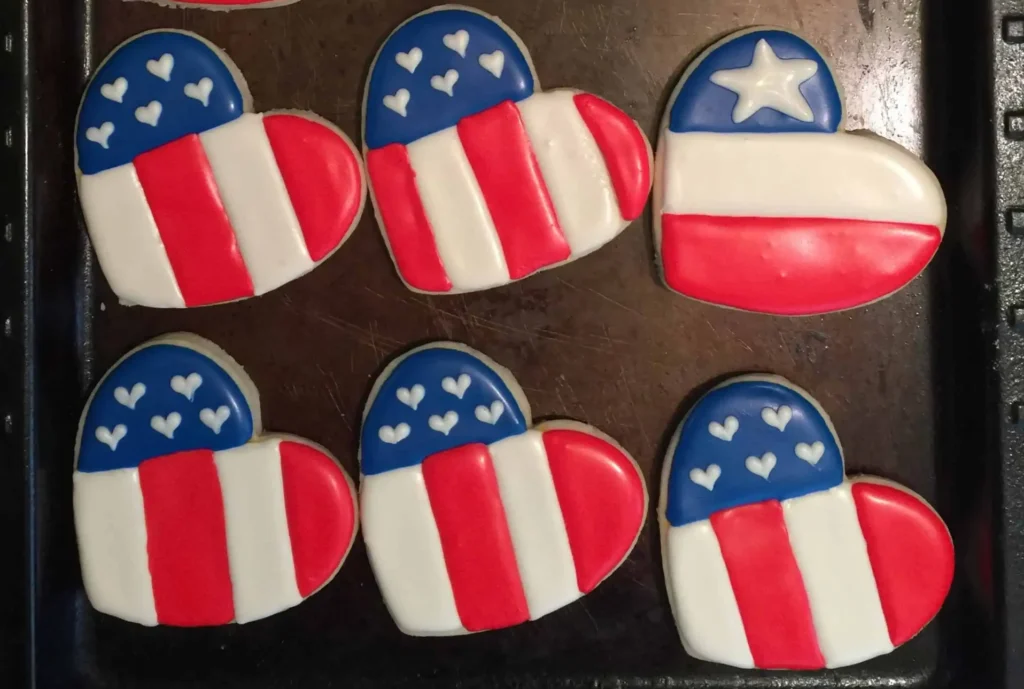 A tray of heart-shaped patriotic cookies decorated with red, white, and blue icing. Some feature a single white star on a blue field with stripes, while others have multiple hearts and vertical flag-like stripes, resembling the American flag.