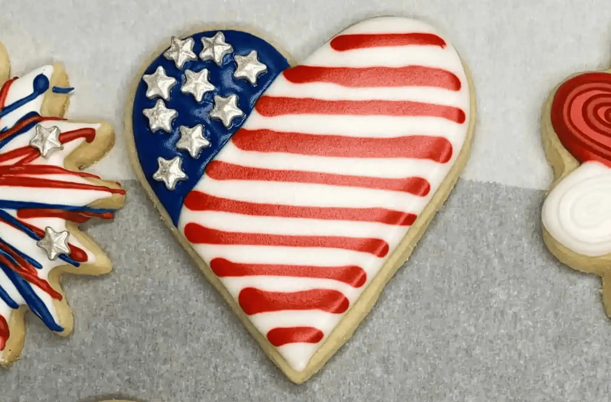 Heart-shaped sugar cookie decorated with red and white icing stripes and a blue section filled with silver stars, styled to resemble the American flag. Perfect for a patriotic sugar cookie tutorial or festive Fourth of July baking blog post.