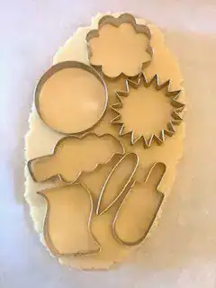 Assortment of cookie cutters pressed into sugar cookie dough, including flower, circle, popsicle, firework, and ice cream shapes.