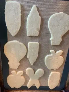 A tray of baked sugar cookies with back-to-school shapes including a pencil, pen, apple, scissors, and hot air balloon, ready for decorating.