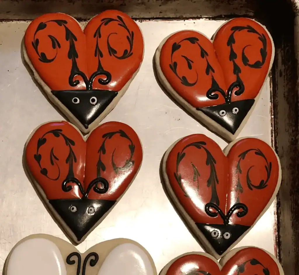 Heart-shaped Halloween sugar cookie decorated with royal icing as a ladybug with a black triangular face, red wings, and whimsical black dots shaped into hearts.