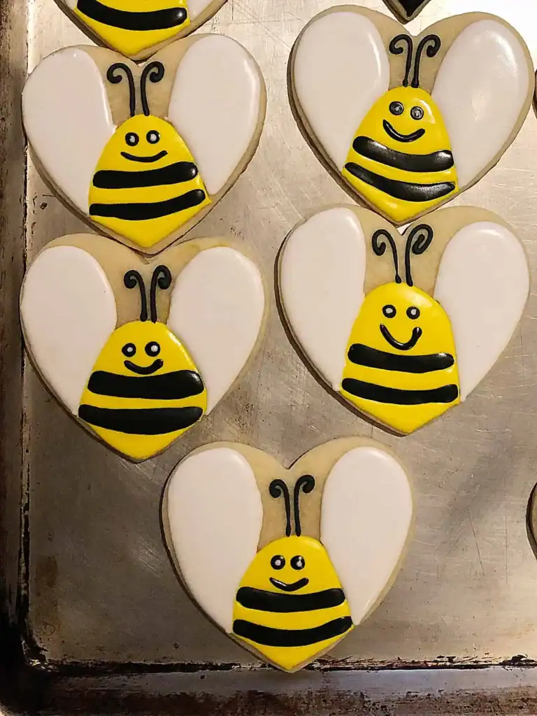 Heart-shaped Halloween sugar cookie decorated with royal icing as a bumble bee with yellow and black stripes, white wings, and a smiley face.