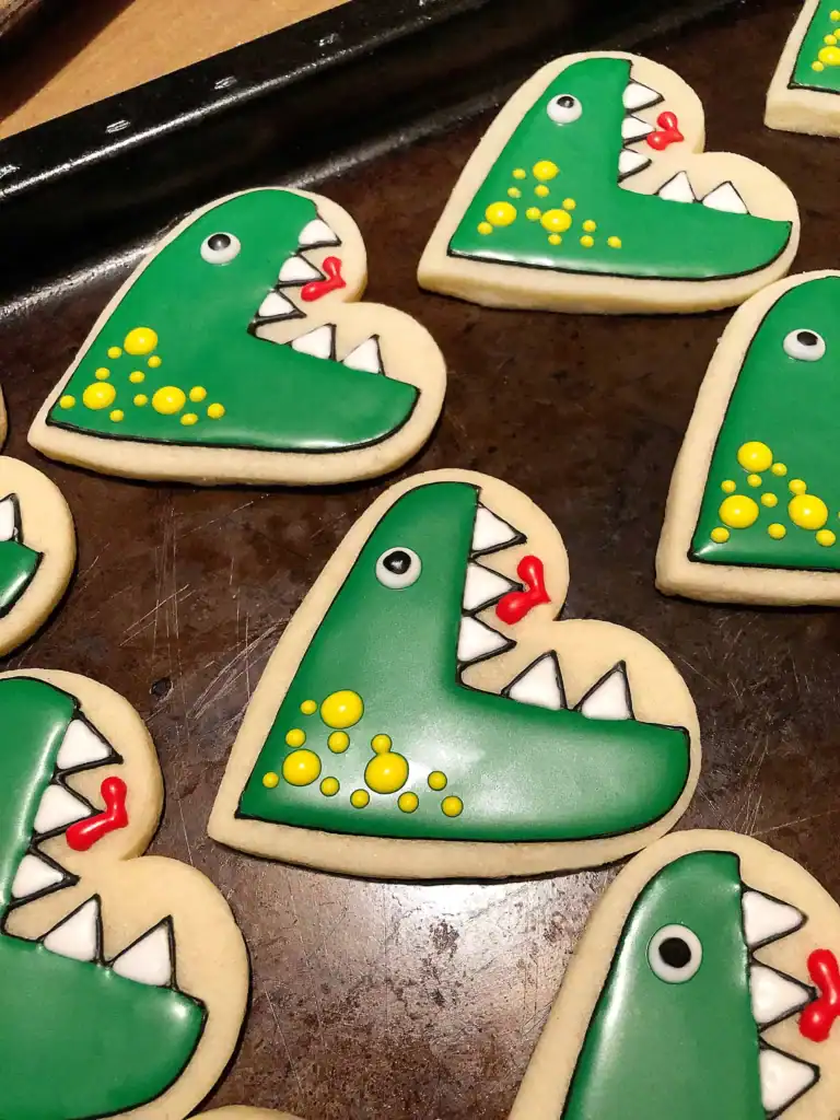 Heart-shaped Halloween sugar cookie decorated with royal icing as a green dinosaur with big white teeth, yellow spots, and a tiny red heart detail.