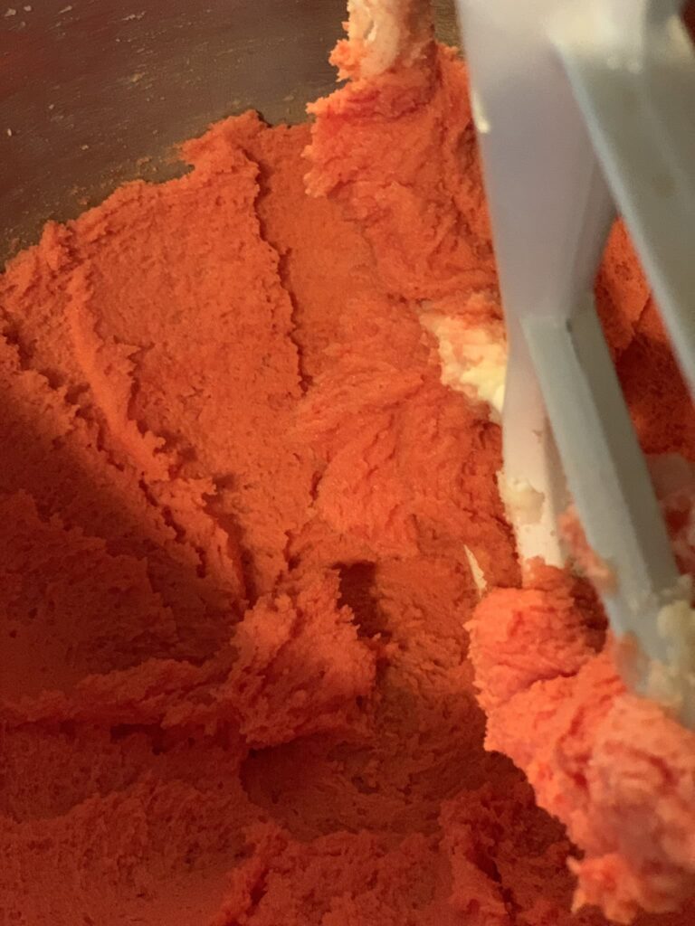 Mixing bowl containing sugar, butter, egg, vanilla, and red food coloring.
