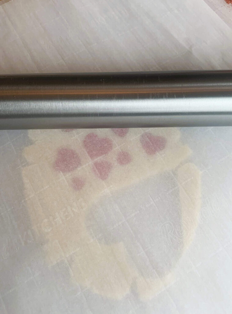 Natural color sugar cookie dough in heart shape with red heart inset pattern between two pieced of parchment paper and lighting rolling over with rolling pin.