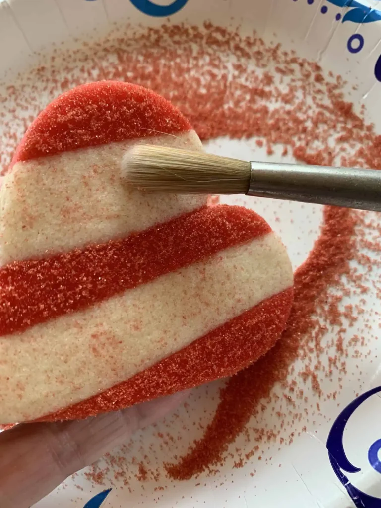 Heart shaped sugar cookie with sanding sugar applied and brushing off excess sanding sugar.
