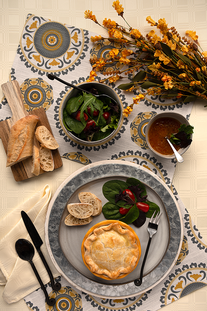 Classic Chicken Pot Pie - Overhead view of a table setting. resting on the table is a salad and dressing, wooden cutting board with baguette sliced, a plate with bread, salad and a mini pot pie.