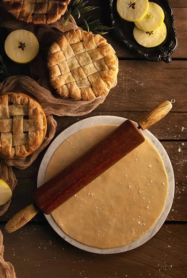 Best Flaky Pie Dough rolled our on a marble surface resting on top is a vintage rolling pin. To the side are three personal sized pies.